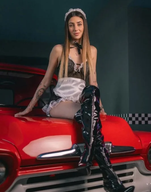 Michelle Comi, modella di OnlyFans, foto seduta sull'auto con un costume da cameriera sexy.